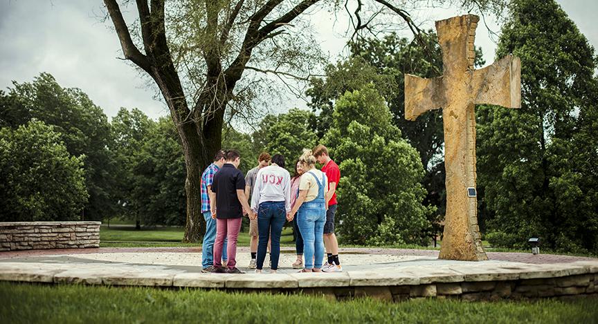 A group of five people stands in a circle holding hands near a large stone cross outdoors, 四周都是树木和绿色植物.